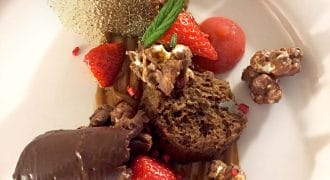 A plated dessert featuring chocolate sponge, chocolate mousse, fresh strawberries, candied nuts, a sugar shard, caramel sauce, and a mint sprig, all artfully arranged on a white plate.