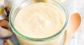 A small glass jar filled with creamy aioli sits on a marble surface, surrounded by garlic cloves, a wooden spoon, a halved lemon, and a white cloth with yellow stripes.