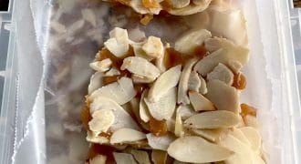Two pieces of almond brittle with caramel and sliced almonds are placed on greaseproof paper inside a rectangular container.