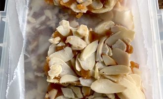 Two pieces of almond brittle with caramel and sliced almonds are placed on greaseproof paper inside a rectangular container.