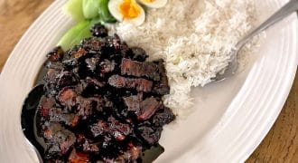 A white oval platter with steamed white rice, soy-braised pork belly in dark sauce, sliced soft-boiled eggs, and blanched green vegetables, served with a large serving spoon.