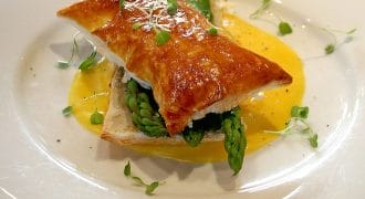 A golden puff pastry sits atop fresh asparagus spears and sauce on a white plate, garnished with microgreens, on a light wooden table. A fork and knife are in the background.
