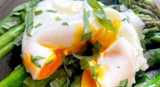 A poached egg with a runny yolk sits atop bright green asparagus spears, garnished with chopped fresh herbs, on a dark plate.