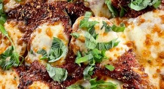 Close-up of baked chicken parmigiana topped with melted mozzarella cheese, tomato sauce, grated parmesan, and fresh basil leaves. The dish has a golden, bubbly, and slightly browned appearance.