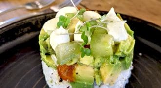 A round stack of sushi rice topped with diced avocado, kiwi, cherry tomatoes, microgreens, and a drizzle of creamy sauce, served on a black plate with a fork and knife in the background.
