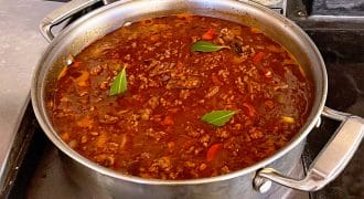 A large stainless steel saucepan on a hob contains simmering meat sauce with visible minced beef, tomato pieces, and bay leaves floating on the surface.