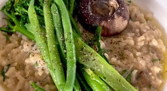 A bowl of creamy risotto topped with steamed tenderstem broccoli and a roasted mushroom, garnished with cracked black pepper and fresh herbs.