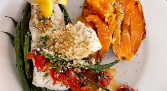 A hand squeezes a lemon over a plate with baked fish topped with breadcrumbs and cherry tomatoes, roasted green beans, and a split baked sweet potato sprinkled with seasoning.