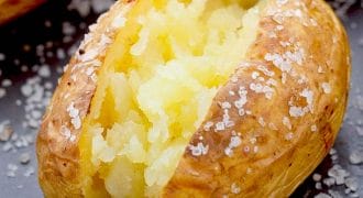 A baked potato split open to reveal its fluffy interior, with coarse salt sprinkled on its crispy golden skin and on the surrounding dark surface.
