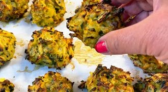 A hand with pink nail varnish picks up a golden-brown baked vegetable fritter from a tray lined with baking paper, surrounded by several similar fritters.