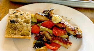 A white plate with roasted vegetables, sausage, and a slice of olive focaccia bread. A baking tray with more roasted vegetables and sausages is in the background on a wooden table.