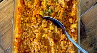 A glass dish filled with creamy orange risotto containing visible pieces of vegetables, such as carrots and broccoli, with a silver spoon resting inside. The dish is placed on a wooden surface.