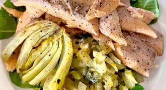 A plate of poached fish fillets topped with black pepper, served on a bed of sautéed greens, onions, and a wedge of cooked artichoke. The dish is arranged on a white plate.