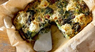 A baked vegetable and cheese frittata in a parchment-lined dish, with a serving spatula and a wedge already removed, sits on a wooden table.