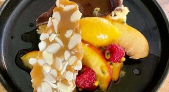 A black plate with a slice of cheesecake, topped with caramel almond brittle, fresh peach slices, raspberries, passion fruit, and drizzled with a dark sauce. The dish is served on a wooden table.