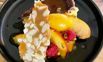 A black plate with a slice of cheesecake, topped with caramel almond brittle, fresh peach slices, raspberries, passion fruit, and drizzled with a dark sauce. The dish is served on a wooden table.