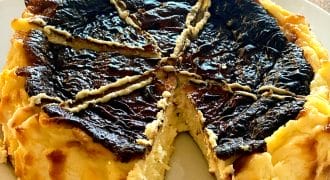 A round, rustic cheesecake with a deeply browned, cracked top sits on a white plate. One large wedge is missing, revealing the creamy interior. The dessert is on a wooden surface.