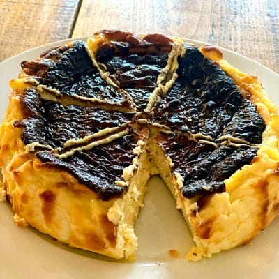 A round, rustic cheesecake with a deeply browned, cracked top sits on a white plate. One large wedge is missing, revealing the creamy interior. The dessert is on a wooden surface.