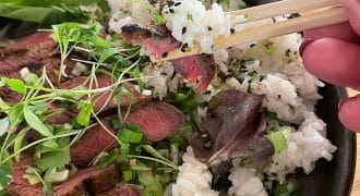 A hand uses chopsticks to pick up a bite of sliced steak and rice from a bowl filled with white rice, sesame seeds, sliced steak, fresh greens, and microgreens.