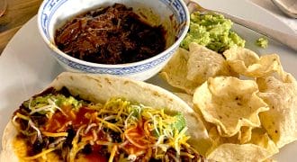 A plate with a soft taco filled with shredded beef, cheese, and sauce, alongside tortilla chips, guacamole, and a bowl of shredded beef. A hot sauce bottle is in the background.