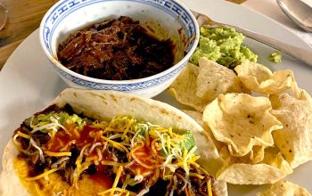 A plate with a soft taco filled with shredded beef, cheese, and sauce, alongside tortilla chips, guacamole, and a bowl of shredded beef. A hot sauce bottle is in the background.