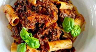 A white bowl filled with rigatoni pasta topped with a rich, chunky meat sauce, garnished with fresh basil leaves.