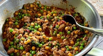 A metal pan filled with a colourful dish of minced meat, green peas, and chickpeas in a tomato-based sauce sits on a wooden table, with a spoon inside. Plates of rice and flatbread are visible in the background.
