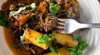 A white plate with rigatoni pasta topped with shredded beef in a rich sauce, diced vegetables, and fresh basil leaves. A fork rests on the plate.