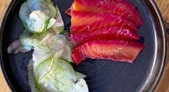 A black plate with slices of beet-cured salmon and a side salad of thinly sliced cucumber, radish, cauliflower, and fresh dill, set on a wooden table.