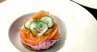 An elegantly plated starter featuring a round base of dark bread topped with a pink creamy spread, slices of smoked salmon, thin cucumber slices, and a sprig of dill, served on a white plate.