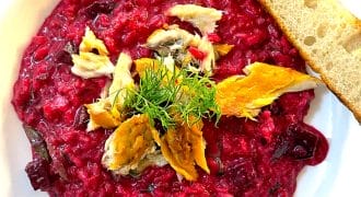 A white bowl of vibrant beetroot risotto, topped with chunks of smoked fish and fresh dill, served with a slice of toasted bread on the side.