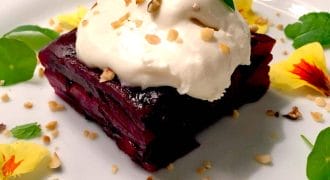 A square slice of beetroot terrine topped with a dollop of cream and chopped nuts, garnished with yellow edible flowers and fresh mint leaves on a white plate.
