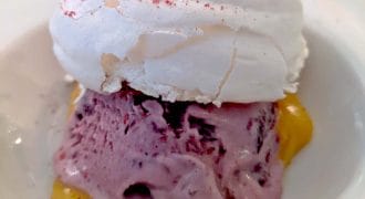 A dessert featuring a scoop of purple berry ice cream on a yellow custard base, topped with a round, crisp white meringue and a sprinkle of pink powder, served in a white bowl.