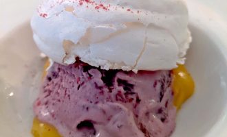 A dessert featuring a scoop of purple berry ice cream on a yellow custard base, topped with a round, crisp white meringue and a sprinkle of pink powder, served in a white bowl.