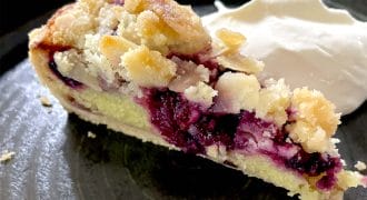 A slice of berry crumble pie with a crumbly topping sits on a dark plate next to a dollop of whipped cream.