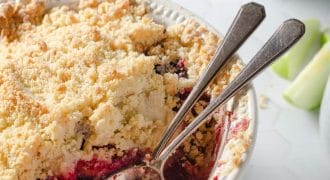 A round baking dish filled with berry crumble, topped with a golden, crumbly crust. Two spoons rest inside, and some fruit filling is visible. In the background, a bowl of berries and apple slices are partially seen.