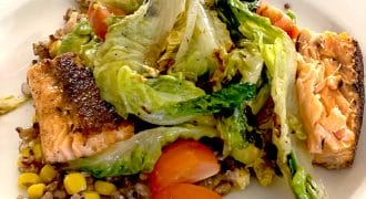 A plate of grilled salmon served with mixed grains, sweetcorn, cherry tomatoes, and sautéed greens, arranged on a white dish.