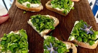 Sliced bread topped with creamy cheese, chopped green peas, herbs, and purple edible flowers arranged on a wooden board, with books visible in the background.