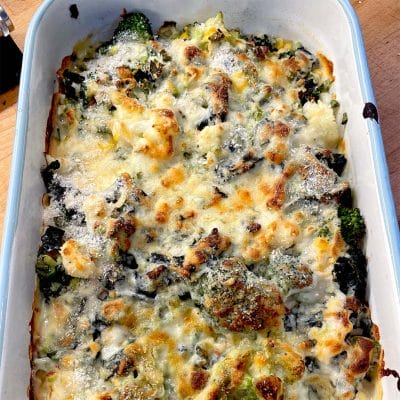 A baked casserole dish filled with broccoli, cauliflower, and melted cheese, browned on top, sitting on a wooden surface.