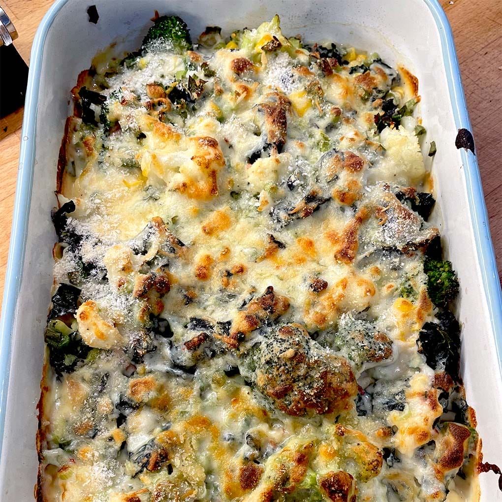 A baked casserole dish filled with broccoli, cauliflower, and melted cheese, browned on top, sitting on a wooden surface.