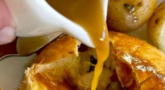 Golden brown gravy is being poured from a small jug over a flaky pastry dish, with roast potatoes visible in the background. The pastry is cracked open, revealing its filling inside.