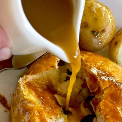 Golden brown gravy is being poured from a small jug over a flaky pastry dish, with roast potatoes visible in the background. The pastry is cracked open, revealing its filling inside.
