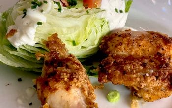 A plate with a wedge of iceberg lettuce topped with creamy dressing, chopped chives, and tomato, served alongside two pieces of crispy fried chicken.