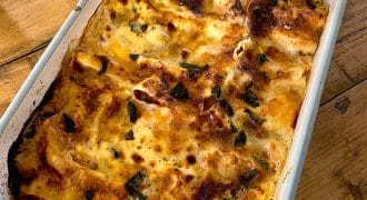 A baked dish of golden-brown lasagne with melted cheese and herbs sits in a white rectangular baking dish on a wooden table.