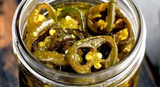 A close-up of a glass jar filled with sliced pickled jalapeños in brine, showing seeds and pepper rings. The jar sits on a dark wooden surface.