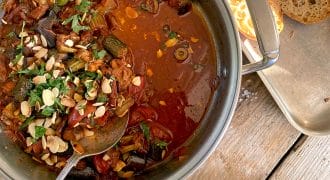 A pot of vegetable stew topped with fresh herbs and sliced almonds sits on a wooden table next to a tray of sliced bread and a plate with green beans.