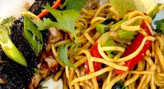 Grilled fish fillet topped with herbs, served alongside stir-fried noodles with red peppers, spring onions, and coriander, garnished with a lime wedge on a white plate.