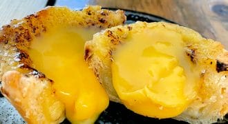 A close-up of a pastry cut in half, revealing a rich, yellow, creamy filling oozing out onto a dark ceramic plate. The pastry has a golden, flaky crust and sits on a wooden table.