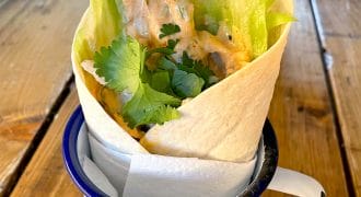A tortilla wrap filled with lettuce, chicken goujons, creamy sauce, and fresh coriander, served upright in a white enamel mug on a wooden table.