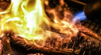 Slices of fish being seared with an open flame, creating a dramatic burst of yellow and orange fire over the surface, highlighting the texture and cooking process in a close-up shot.